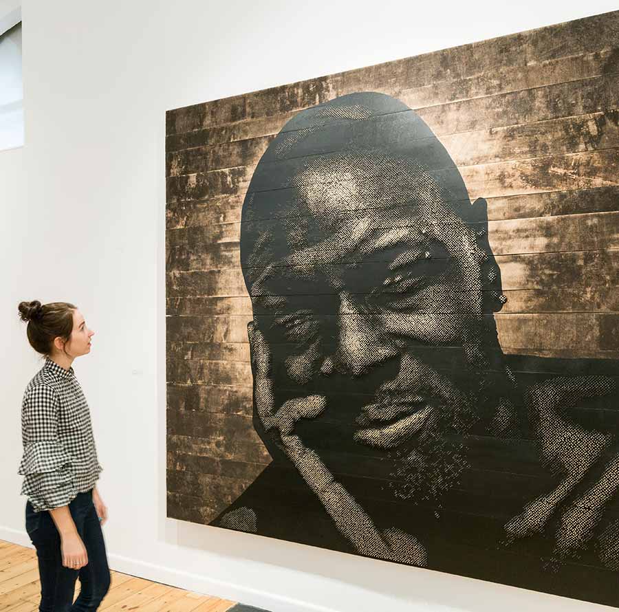 Dee Haughney, Curator, October Gallery, in front of Alexis Peskine, The Architects of New Djenné, 2017. Moon gold leaf on nails, earth, coffee, water and acrylic on wood, 240 x 250 cm, installed at October Gallery. Photo: © Jonathan Greet.